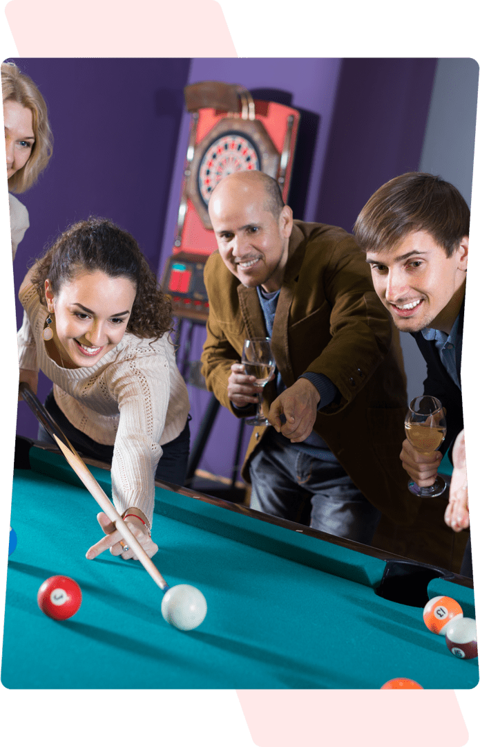 Group of four people playing pool indoors, with two men and two women. One woman is taking a shot while the others watch and hold drinks. Dartboard visible in the background.
