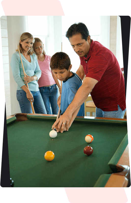 A man instructs a boy on how to play pool while two women observe in the background. There are colored balls on the pool table.