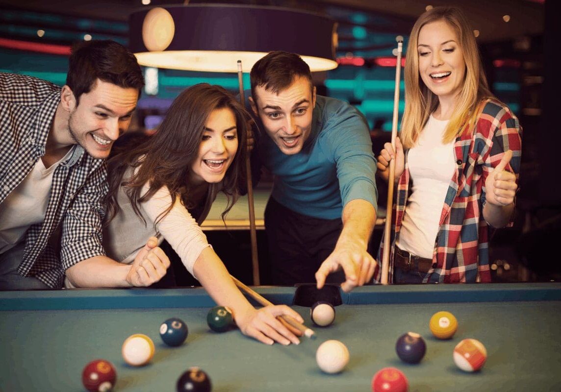 Group of four people playing pool indoors, with two men and two women. One woman is taking a shot while the others watch and hold drinks. Dartboard visible in the background.